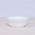 A plain white Re-Play Bowl, crafted from recycled plastic by REPLAY, sits on a light wooden surface against a white tiled backsplash. There are no visible patterns or designs on the bowl, and the background is simple and uncluttered. This piece fits seamlessly into any vibrant tableware set and is safe for kids.