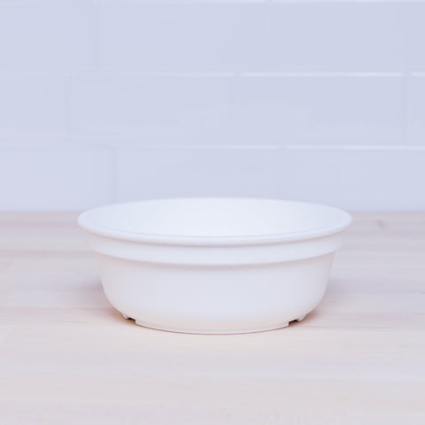 A plain white Re-Play Bowl, crafted from recycled plastic by REPLAY, sits on a light wooden surface against a white tiled backsplash. There are no visible patterns or designs on the bowl, and the background is simple and uncluttered. This piece fits seamlessly into any vibrant tableware set and is safe for kids.