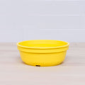 A bright yellow Re-Play Bowl from REPLAY's vibrant tableware set is placed on a light wooden surface with a white tiled background. The round bowl, made from recycled plastic and safe for kids, is empty and features a simple, smooth design.