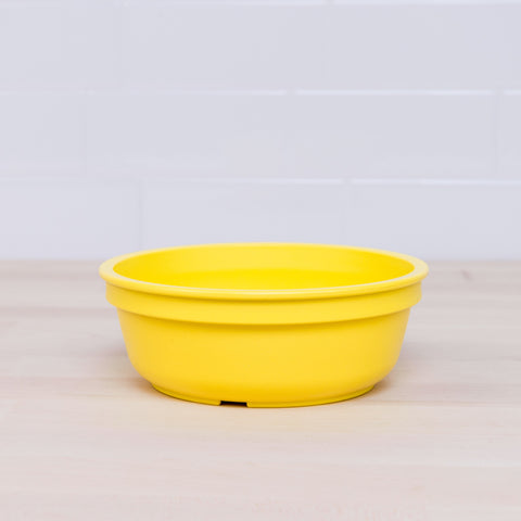 A bright yellow Re-Play Bowl from REPLAY's vibrant tableware set is placed on a light wooden surface with a white tiled background. The round bowl, made from recycled plastic and safe for kids, is empty and features a simple, smooth design.