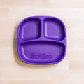 A REPLAY Re-Play Divided Plate in purple, made from durable recycled plastic, is placed on a light wooden surface. Designed as a toddler plate for portion control and organizing various food items, it features one large compartment and two smaller ones.