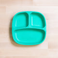 A REPLAY Re-Play Divided Plate, teal and rectangular, crafted from recycled plastic, rests on a light wooden surface. The toddler plate showcases two smaller compartments at the top and one larger compartment at the bottom, with "Re-Play" embossed in the center of the portion control plate.