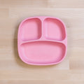 A light pink, square-shaped, Re-Play Divided Plate made from recycled plastic is placed on a light wooden surface. It features three compartments for perfect portion control, with two smaller sections at the top and a larger section at the bottom. The brand name "REPLAY" is embossed near the lower edge.