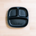 A black Re-Play Divided Plate by REPLAY rests on a light wooden surface. Crafted from recycled plastic, this plate offers three compartments for portion control: one large section at the bottom and two smaller sections at the top. The larger compartment is embossed with the word "Re-Play," making it ideal for use as toddler plates.