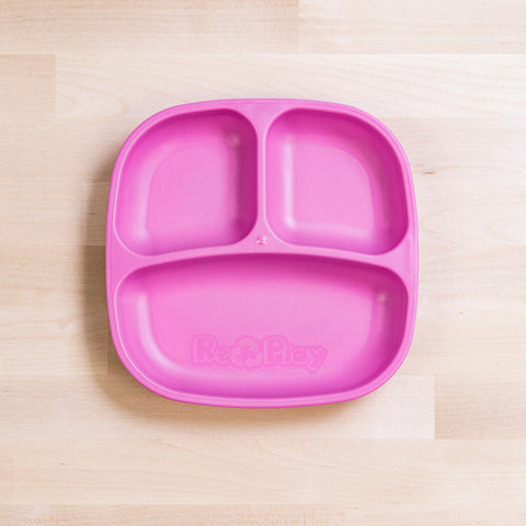 A bright pink Re-Play Divided Plate made by REPLAY, featuring three compartments perfect for portioning various foods, is placed on a light wood surface. Made from recycled plastic plates, it proudly displays the "Re-Play" logo embossed at the bottom.