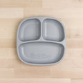 The Re-Play Divided Plate by REPLAY is a gray rectangular portion control plate with three compartments, perfect for serving separate portions of food. Made from recycled plastic, this toddler plate rests on a light wooden surface. The center features the "Re-Play" logo.