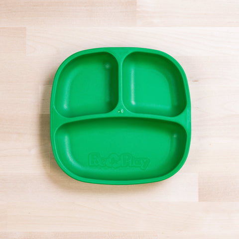 A green Re-Play Divided Plate by REPLAY, made from recycled plastic, sits on a light wooden surface. The plate features three compartments with two smaller sections at the top and a larger one spanning the bottom. "Re-Play" is embossed at the center, making it ideal for portion control.