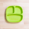 A green REPLAY Re-Play Divided Plate made from recycled plastic rests on a light wooden surface. It features two smaller compartments on the top side and one larger compartment on the bottom side, making it perfect for portion control. The brand name "REPLAY" is embossed on the plate.