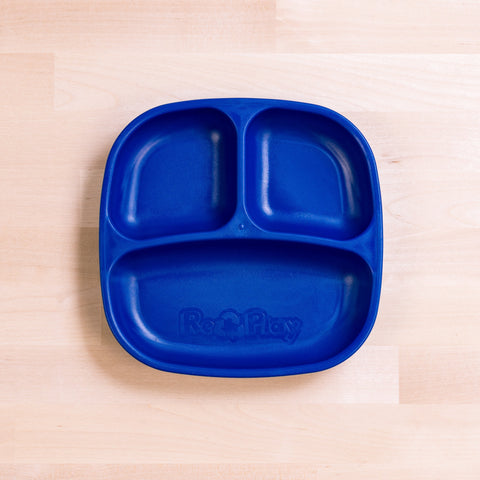 The Re-Play Divided Plate is a blue toddler plate crafted from recycled plastic, featuring three compartments for portion control. It includes one large section at the bottom and two smaller sections at the top. The plate has a slightly raised edge and displays the brand name "REPLAY" embossed in the large section, resting on a light wooden surface.