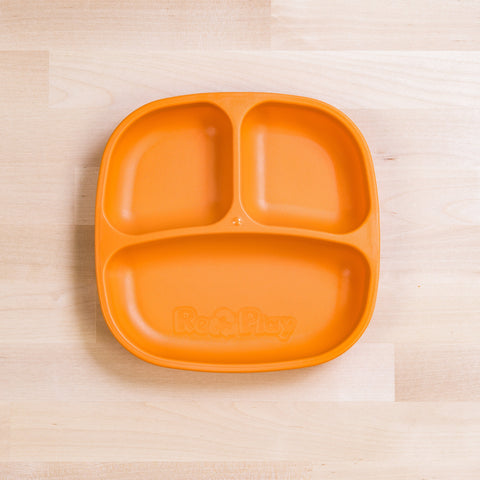 A Re-Play Divided Plate from the brand REPLAY, with three compartments, is resting on a light wooden surface. The orange toddler plate has "Re-Play" embossed on its bottom compartment. Made from recycled plastic, it boasts a smooth finish with varying light wood grain patterns visible underneath.