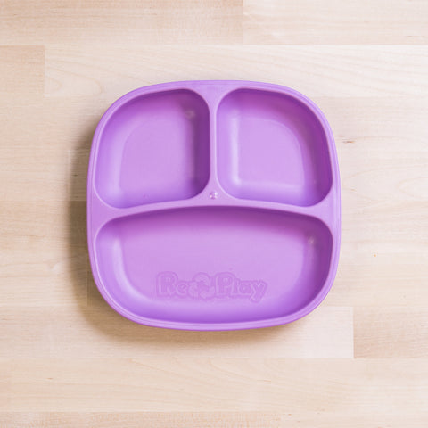 The Re-Play Divided Plate in a vibrant purple color is sitting on a light wooden surface. This portion control plate, made from recycled plastic, features three compartments: a larger one at the bottom and two smaller ones at the top. The brand name "REPLAY" is prominently embossed on the lower section of the plate.