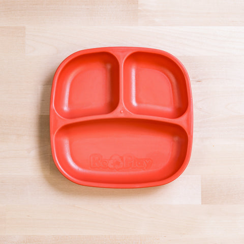 The REPLAY Re-Play Divided Plate is a bright red recycled plastic children's plate featuring three sections, one larger at the bottom and two smaller at the top. The "Re-Play" logo is embossed on the bottom section. Ideal for portion control, this toddler plate is placed on a light wooden surface.