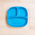 A Re-Play Divided Plate from REPLAY, made of blue recycled plastic and featuring three compartments, sits on a light wooden surface. The portion control plate is designed with one large section and two smaller sections to efficiently portion different foods.