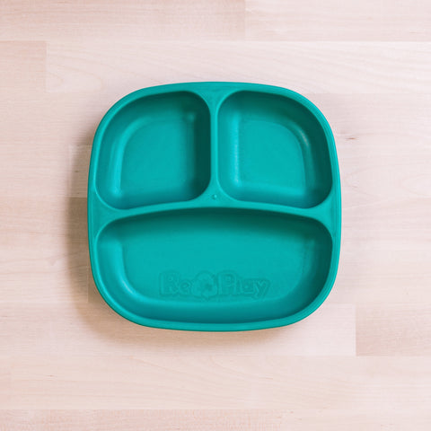 A teal REPLAY Re-Play Divided Plate, designed with three sections and a recycling theme, rests on a light wood surface. Constructed from recycled plastic, it features two smaller top sections and one larger bottom section. The word "Re-Play" is embossed on the bottom section, ideal for encouraging eco-friendly meal times for toddlers.