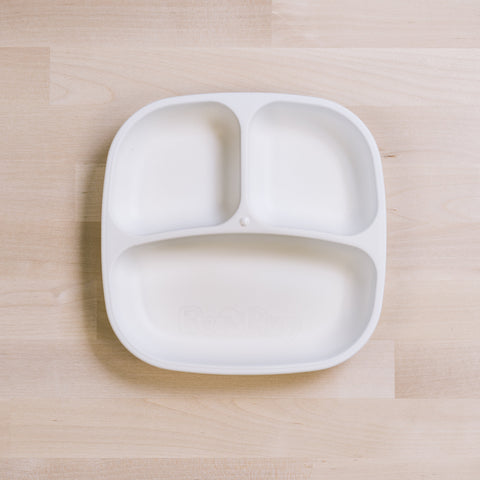 A white Re-Play Divided Plate, ideal for portion control, sits on a light wood surface. Made from recycled plastic by the brand REPLAY, the plate features two smaller compartments on the top and one larger compartment on the bottom, perfect for toddlers.