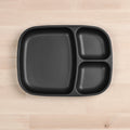 The Re-Play Divided Tray by REPLAY is a black, rectangular, plastic plate with rounded edges that features three compartments: one large section on the left and two smaller sections stacked vertically on the right. Made from recycled materials, this durable tableware rests elegantly on a light wooden surface.