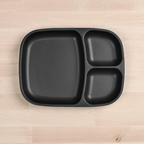 The Re-Play Divided Tray by REPLAY is a black, rectangular, plastic plate with rounded edges that features three compartments: one large section on the left and two smaller sections stacked vertically on the right. Made from recycled materials, this durable tableware rests elegantly on a light wooden surface.