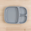 The REPLAY Re-Play Divided Tray is a gray plastic plate made from recycled materials, featuring four compartments of varying sizes, and it's placed on a light wood surface. The largest compartment is located on the left half, while the right half is divided into three smaller sections. This durable tableware exemplifies smart, eco-friendly design.