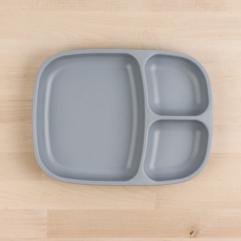 The REPLAY Re-Play Divided Tray is a gray plastic plate made from recycled materials, featuring four compartments of varying sizes, and it's placed on a light wood surface. The largest compartment is located on the left half, while the right half is divided into three smaller sections. This durable tableware exemplifies smart, eco-friendly design.