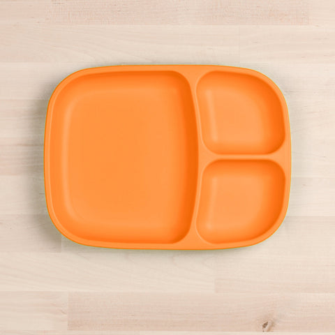 A Re-Play Divided Tray from REPLAY, featuring a bright orange color and made from recycled plastic, is positioned on a light wooden surface. This eco-friendly plate has three compartments: one large section on the left and two smaller sections on the right.