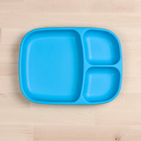 The REPLAY Re-Play Divided Tray, a bright blue serving tray made from recycled plastic, features three compartments: one large section on the left and two smaller sections on the right. Perfect for durable tableware, it sits elegantly on a light wooden surface.