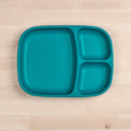 The REPLAY Re-Play Divided Tray, a turquoise plate featuring one large and two smaller compartments, sits on a light wooden surface. Crafted from recycled material, the tray highlights rounded edges and a smooth texture.