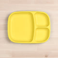 A bright yellow Re-Play Divided Tray made by REPLAY, crafted from recycled materials and featuring three sections, lies on a light wooden surface. One section is larger and rectangular, while the other two are smaller and square-shaped. The durable tableware is empty and clean.