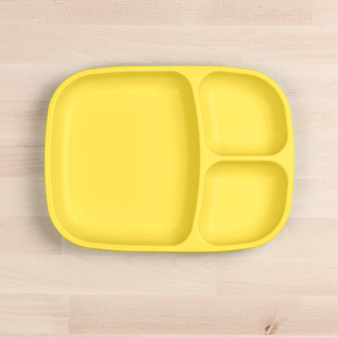 A bright yellow Re-Play Divided Tray made by REPLAY, crafted from recycled materials and featuring three sections, lies on a light wooden surface. One section is larger and rectangular, while the other two are smaller and square-shaped. The durable tableware is empty and clean.