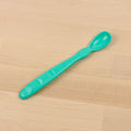 A Re-Play Infant Spoon by REPLAY, featuring a plastic teal-colored design made from recycled material, lies on a light wood surface. It has a curved handle with a series of indentations for grip and a small, rounded bowl.