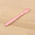 The Re-Play Infant Spoon from REPLAY lies flat on a wooden surface. Crafted from recycled materials, the pink spoon features a rounded handle with subtle indentations for grip and a small, shallow bowl suitable for feeding infants. The wooden background has a light, natural finish that perfectly complements this sustainable tableware piece.