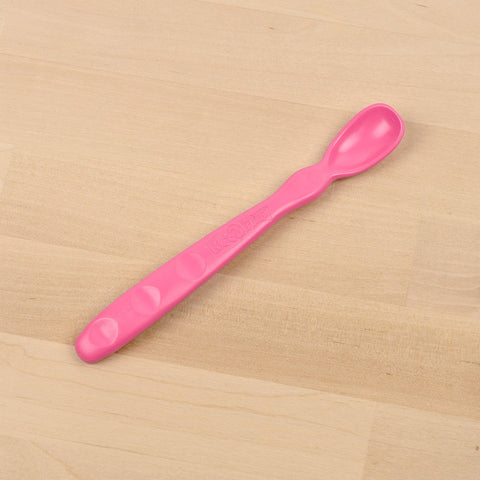 A Re-Play Infant Spoon by REPLAY, featuring a pink plastic design with a long handle and rounded tip, lies on a light wooden surface. The handle includes small round indents for grip and has the word "Bébé" embossed on it. Made from recycled materials, this sustainable tableware ensures both safety and eco-friendliness.