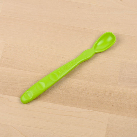 A Re-Play Infant Spoon from REPLAY, crafted in green from recycled materials, featuring a textured handle, lays flat on a light wooden surface. This sustainable tableware item has an elongated, smooth bowl and tactile bumps on the handle for a better grip.