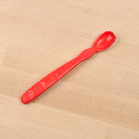 The REPLAY Re-Play Infant Spoon is a red, plastic baby spoon with a textured handle and a rounded, shallow bowl tip made from recycled material. This sustainable tableware item is placed on a light wooden surface.