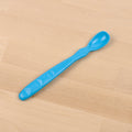 A REPLAY Re-Play Infant Spoon, featuring a blue plastic curved handle and a small oval-shaped bowl made from recycled material, rests on a light wooden surface.