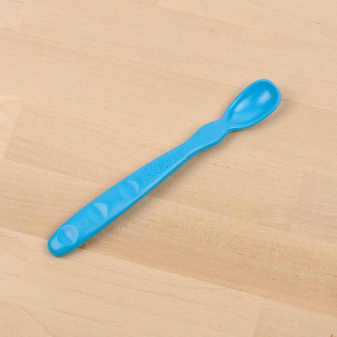 A REPLAY Re-Play Infant Spoon, featuring a blue plastic curved handle and a small oval-shaped bowl made from recycled material, rests on a light wooden surface.