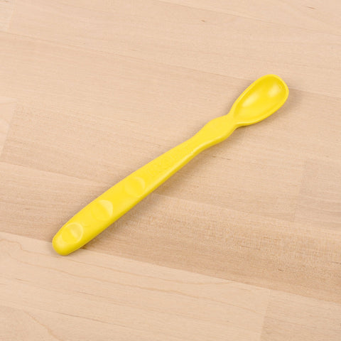 The Re-Play Infant Spoon, in a bright yellow hue, rests on a light wood surface. Crafted from recycled materials by the brand REPLAY, its ergonomic handle features contoured indents for an easy grip, while the small, shallow bowl is ideal for feeding infants. This sustainable tableware choice ensures both functionality and eco-friendliness.