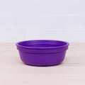 A Re-Play Large Bowl by REPLAY in purple, made from BPA-free plastic, sits on a light wooden surface in front of a white tiled wall. The round bowl with slightly raised edges is perfect for sustainable tableware collections.