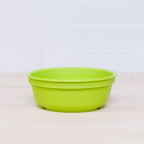 The Re-Play Large Bowl by REPLAY, a bright green shallow bowl made from BPA-free recycled plastic, is placed on a light wooden surface with a white tiled background. The empty bowl highlights the minimalistic and sustainable tableware setup.