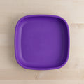 The Re-Play Large Flat Plate by REPLAY, a bright purple, square-shaped plate with slightly rounded edges, rests on a light wooden surface. Crafted from recycled plastic dinnerware, the plate's simple and smooth design is unadorned.