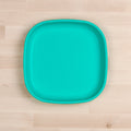 A turquoise Re-Play Large Flat Plate by REPLAY with rounded edges lies on a light wooden surface. Made in America, the plate features a simple, modern design with smooth curves and a solid color, making it an attractive and versatile piece for various table settings.