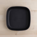 The REPLAY Re-Play Large Flat Plate, characterized by its black square shape with softly rounded edges, is presented on a light wood surface. Crafted in America from recycled plastic dinnerware, the plate boasts a smooth, matte finish that exudes a minimalist and modern charm.