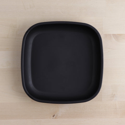 The REPLAY Re-Play Large Flat Plate, characterized by its black square shape with softly rounded edges, is presented on a light wood surface. Crafted in America from recycled plastic dinnerware, the plate boasts a smooth, matte finish that exudes a minimalist and modern charm.