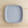 A top-down view of a Re-Play Large Flat Plate from REPLAY, showcasing its plain, light gray, square-shaped design with rounded edges on a light wooden surface. Made in America from recycled plastic dinnerware, the BPA-free plate is empty and smooth, featuring no visible patterns or designs.