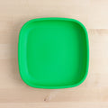 The Re-Play Large Flat Plate by REPLAY, a square, bright green plastic plate with rounded corners made from recycled dinnerware, is placed on a light wood surface. The plate is empty and positioned in the center of the image.