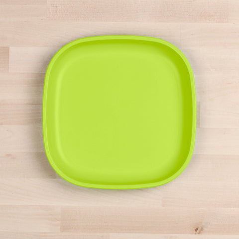 A Re-Play Large Flat Plate, a square green plate with rounded edges made from recycled plastic dinnerware by REPLAY, is placed on a light wooden surface. The plate is empty and centrally positioned in the image.