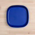 A "Re-Play Large Flat Plate" from REPLAY, featuring a square shape with rounded edges in blue and made from recycled plastic dinnerware, is placed on a light wooden surface.