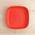 The REPLAY Re-Play Large Flat Plate is a red, square plastic plate with slightly rounded corners, shown on a light wooden surface. Made in America from recycled plastic dinnerware, the plate features a smooth, matte finish and a raised edge around the perimeter. The background highlights the grain and texture of the wooden surface.