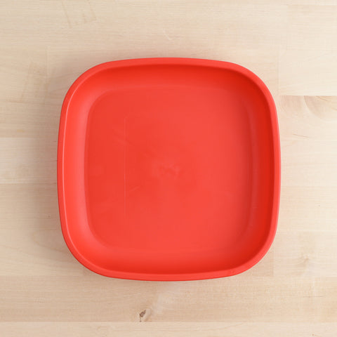 The REPLAY Re-Play Large Flat Plate is a red, square plastic plate with slightly rounded corners, shown on a light wooden surface. Made in America from recycled plastic dinnerware, the plate features a smooth, matte finish and a raised edge around the perimeter. The background highlights the grain and texture of the wooden surface.