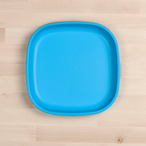 A square, blue REPLAY Re-Play Large Flat Plate made from recycled plastic dinnerware with rounded edges is placed on a light wooden surface. The plate is empty and the background displays a uniform, light wood grain texture.