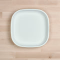 A Re-Play Large Flat Plate from REPLAY, with a white, square design featuring rounded edges, is placed on a light wooden surface. The plate's simple and minimalist aesthetics are free of any visible patterns or decorations. Made in America and BPA-free, the smooth matte finish of the plate contrasts slightly with the wooden background.